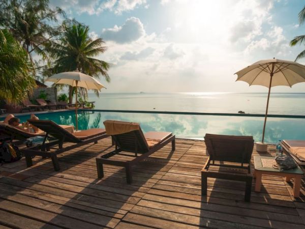 A serene setting with lounge chairs, umbrellas, and a pool overlooking the ocean, surrounded by palm trees under a partly cloudy sky.
