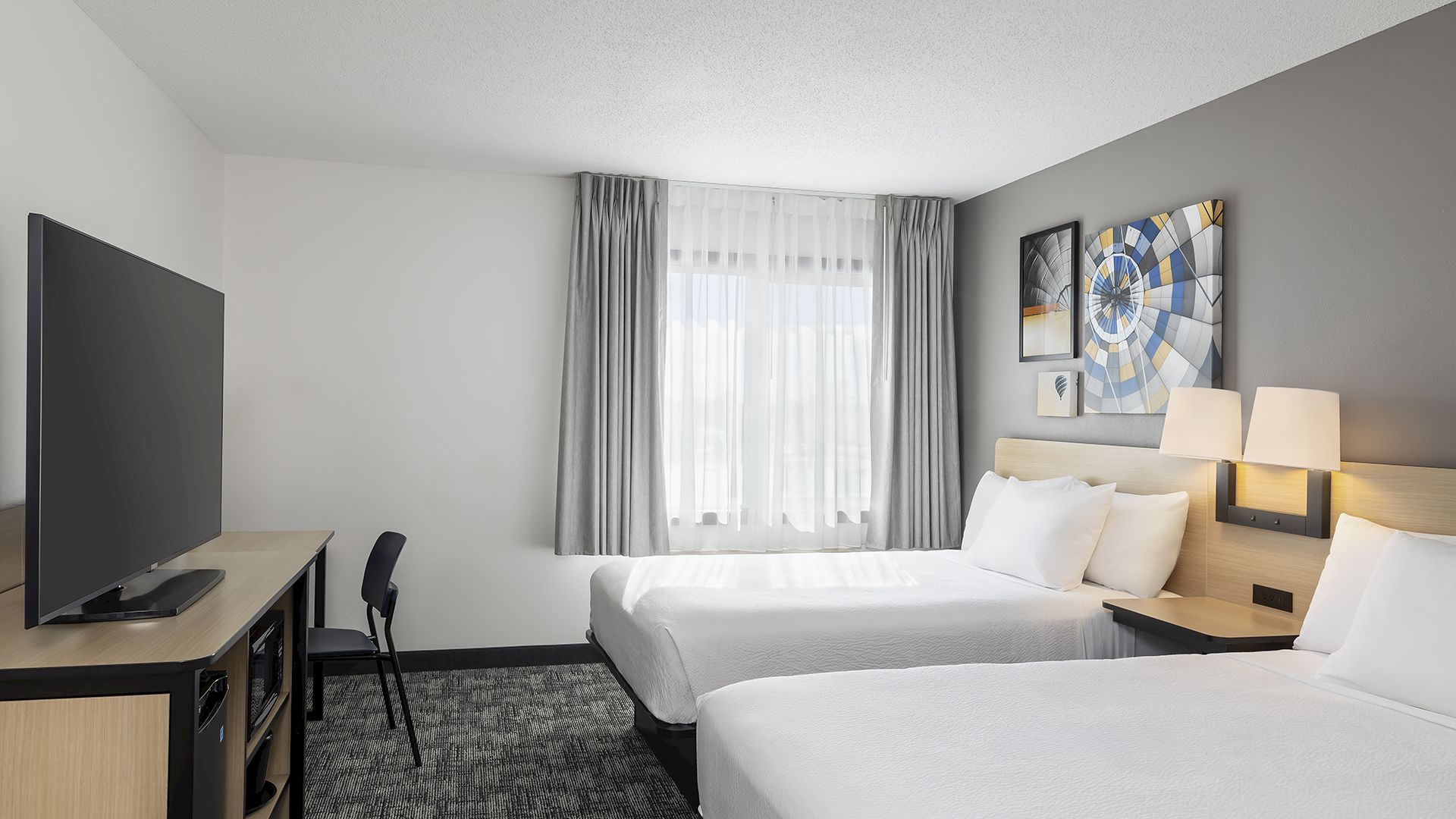A modern hotel room with two double beds, a desk and chair, a flat-screen TV, gray and white decor, and framed wall art by the window. Ends this sentence.