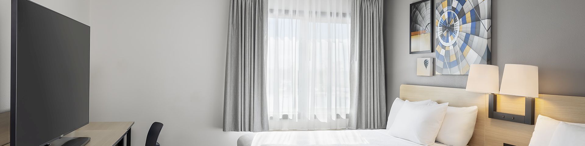 A modern hotel room with two double beds, a desk and chair, a flat-screen TV, gray and white decor, and framed wall art by the window. Ends this sentence.