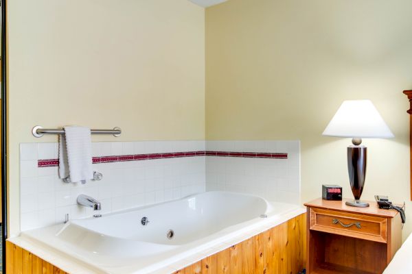 A cozy room with a bathtub, a wooden nightstand next to it, a lamp, and a small alarm clock on a patterned carpeted floor.