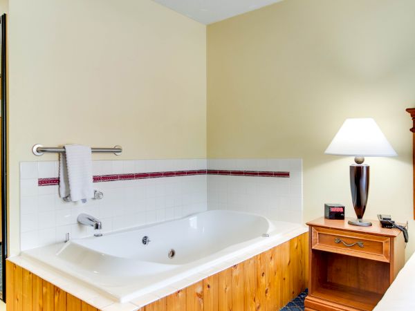 A cozy room with a bathtub, a wooden nightstand next to it, a lamp, and a small alarm clock on a patterned carpeted floor.