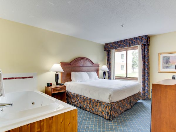 A hotel room with a large bed, bedside tables with lamps, a whirlpool tub, a window with curtains, a framed picture, and a dresser is shown.