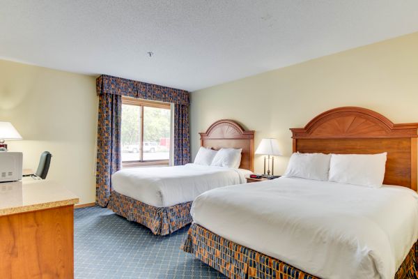 The image shows a hotel room with two double beds, a wooden desk, a chair, lamps, and a window with curtains.