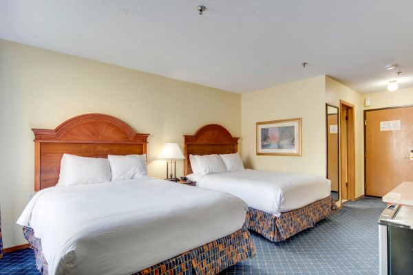 A hotel room with two double beds, matching headboards, white linens, a lamp, framed artwork, and a door leading to the hallway.