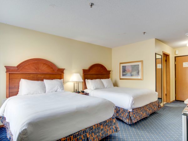 A hotel room with two double beds, matching headboards, white linens, a lamp, framed artwork, and a door leading to the hallway.