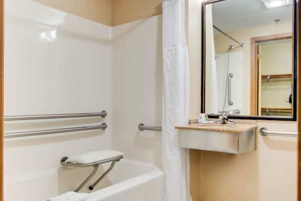 The image shows a bathroom with a bathtub, a safety grab bar, a foldable bench, a sink with a large mirror, and white tiled flooring.