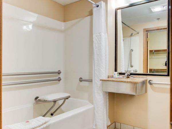 The image shows a bathroom with a bathtub, a safety grab bar, a foldable bench, a sink with a large mirror, and white tiled flooring.