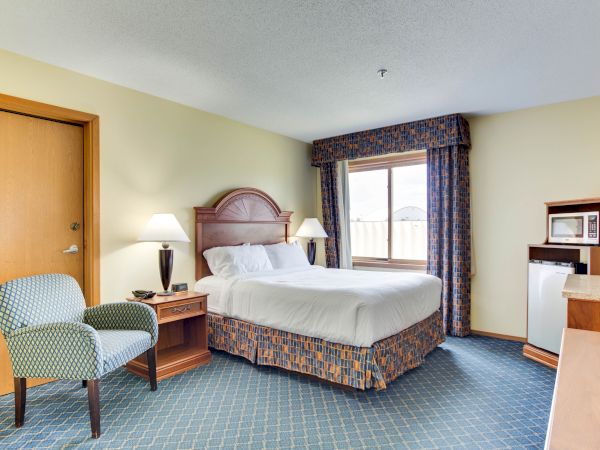 A hotel room with a bed, nightstands with lamps, a green chair, a window with curtains, and a small fridge and microwave on the right side end the sentence.