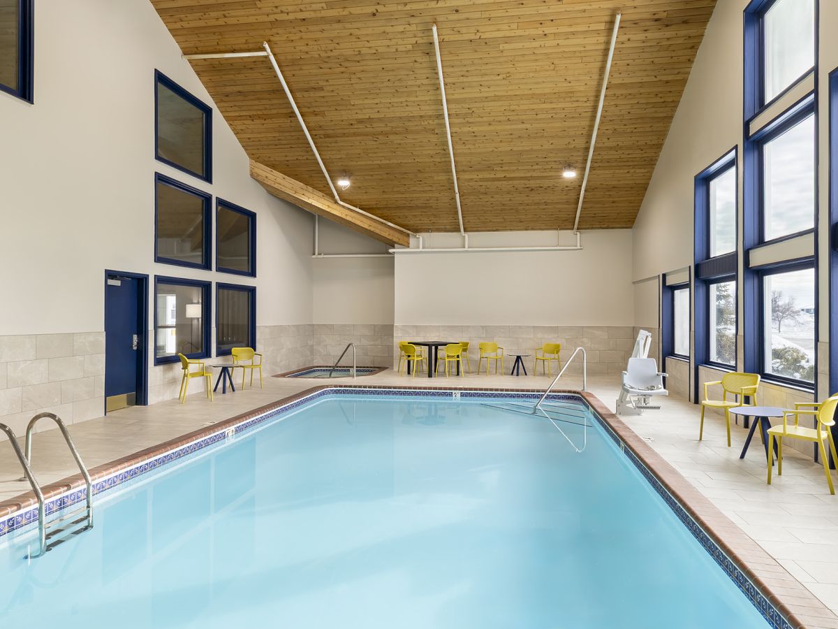 A bright indoor pool area with blue water, beige tiles, and several yellow chairs along the windows, under a wooden ceiling.