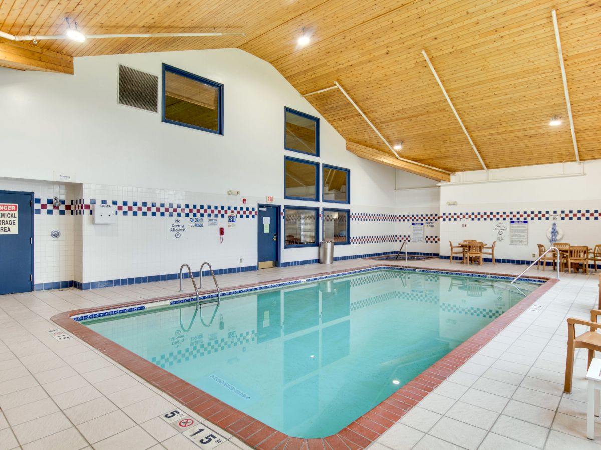 An indoor swimming pool with blue trim, tiled walls, a wooden ceiling, and seating areas with tables and chairs on the surrounding deck.