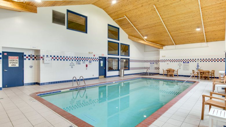 An indoor swimming pool with blue trim, tiled walls, a wooden ceiling, and seating areas with tables and chairs on the surrounding deck.