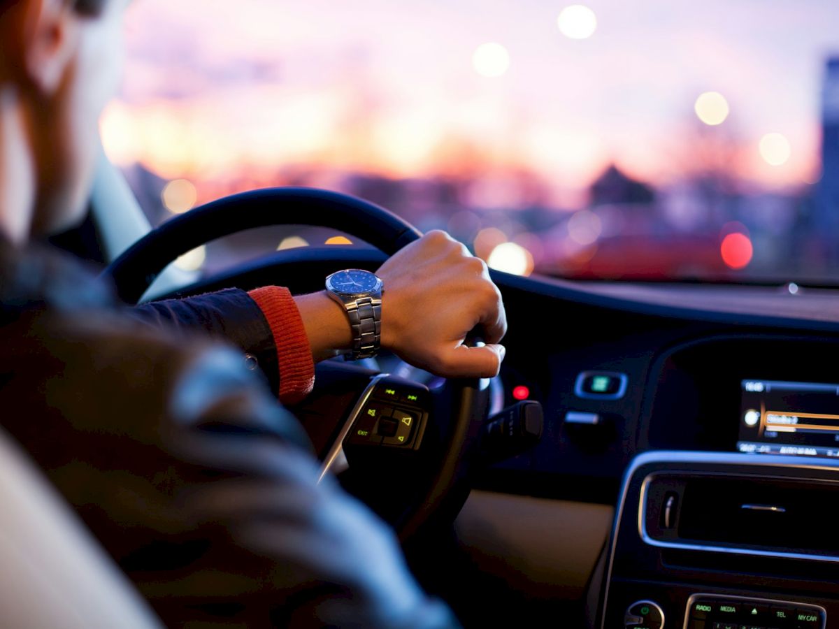 A person is driving a car during sunset with their left hand on the steering wheel and a view of the car's dashboard and instrument panel.