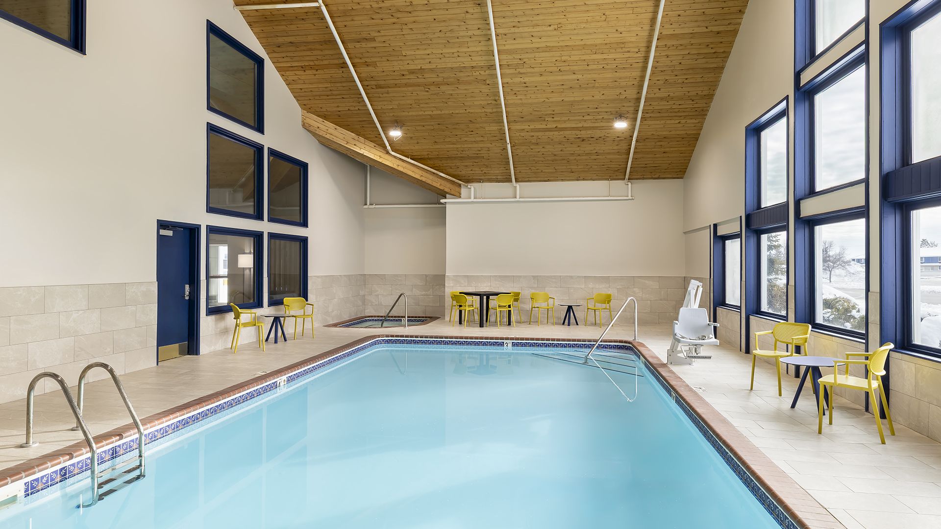 A bright indoor swimming pool area with blue water, tiled floor, large windows, and yellow seating around the edges under a wooden ceiling.