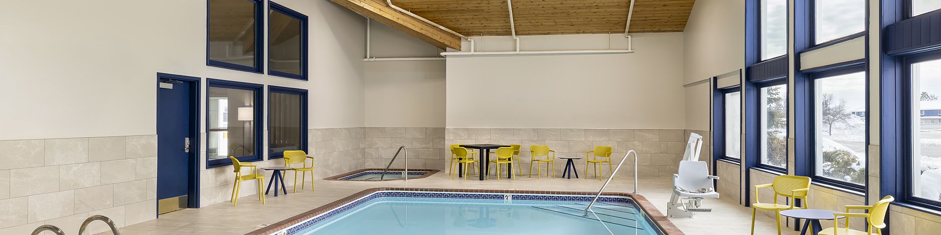 A bright indoor swimming pool area with blue water, tiled floor, large windows, and yellow seating around the edges under a wooden ceiling.