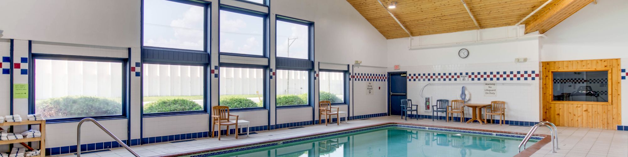 An indoor swimming pool area with large windows, wooden ceiling, and poolside seating. The setting is clean and well-lit, suitable for relaxation.