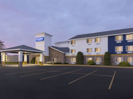 A hotel exterior at dusk with a large parking lot and a covered entrance, modern brick-and-stone facade, and neatly trimmed bushes.