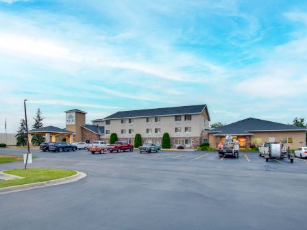 The image shows a parking lot in front of a building, likely a hotel or motel, under a partly cloudy sky with several vehicles parked.