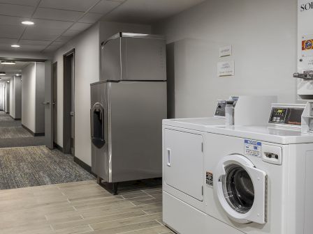 A laundry area in a hotel hallway: a front-loading washing machine, a large industrial dryer, and a hallway leading to guest rooms.