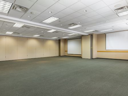 A mostly empty conference room with beige walls, a gray carpet, a few windows/blinds, ceiling lights, and projector screen at the far end, ready for a meeting.