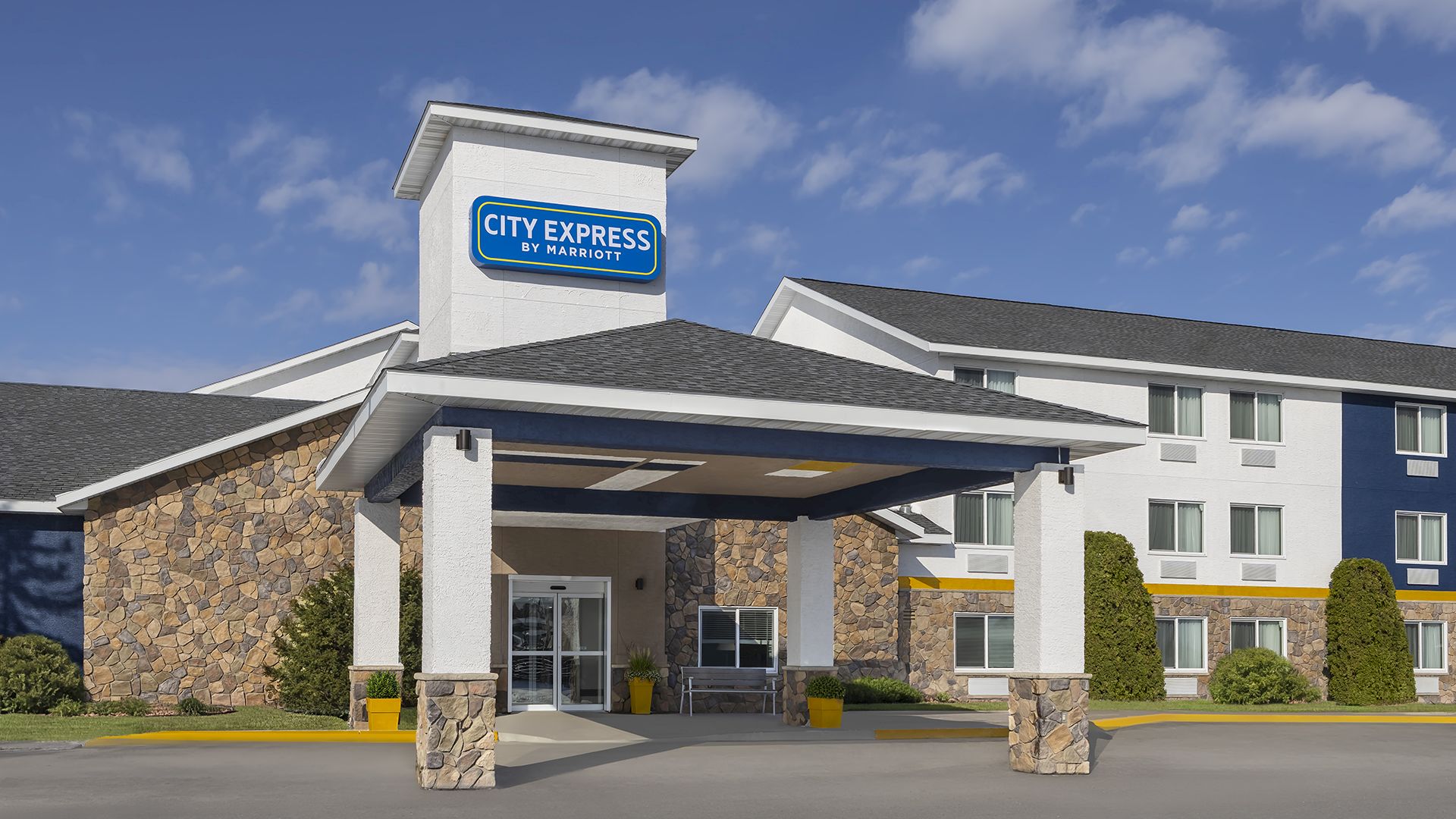 A City Express by Marriott hotel entrance with a &ldquo;CITY EXPRESS&rdquo; sign, stone facade, and trees under a blue sky with clouds.