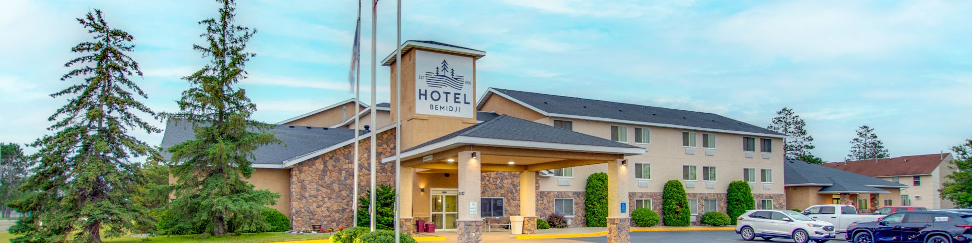 The image shows a hotel building with a parking lot in front, multiple cars parked, a few flags, and a landscaped area.