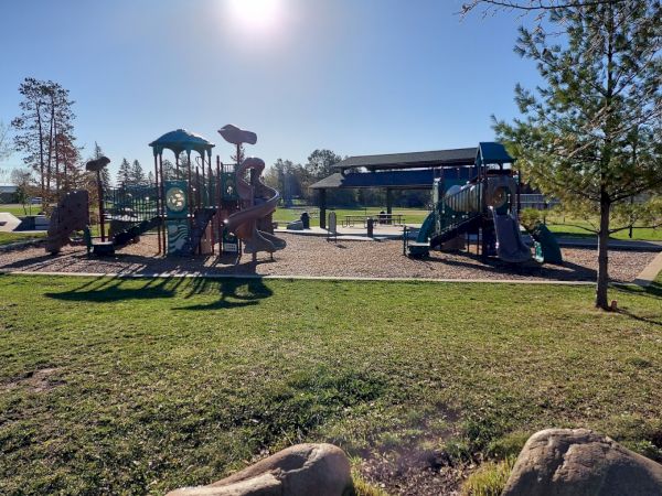 This image shows a playground with slides, climbing structures, and trees, set in a grassy area under a clear, sunny sky.