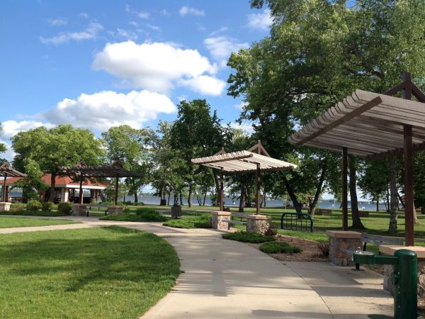 A sunny park with a walking path, green trees, and pavilions under a clear sky with fluffy clouds.
