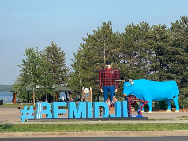 A statue of a lumberjack and a large blue ox stands behind large hashtag "#BEMIDJI" letters, with trees and a lake in the background.