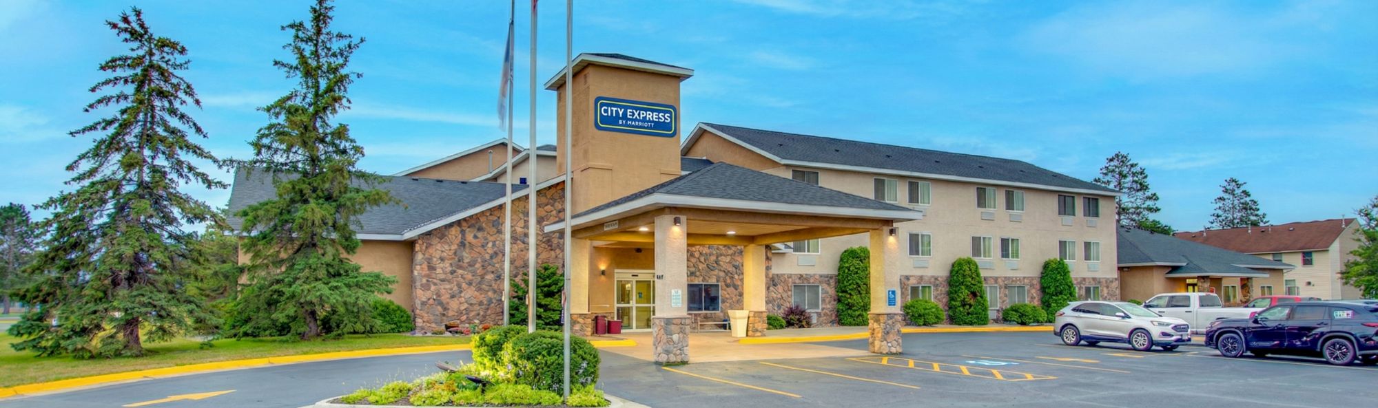 A City Express hotel with a brick and stone facade, a covered entrance, tall trees nearby, and several parked cars in the lot.