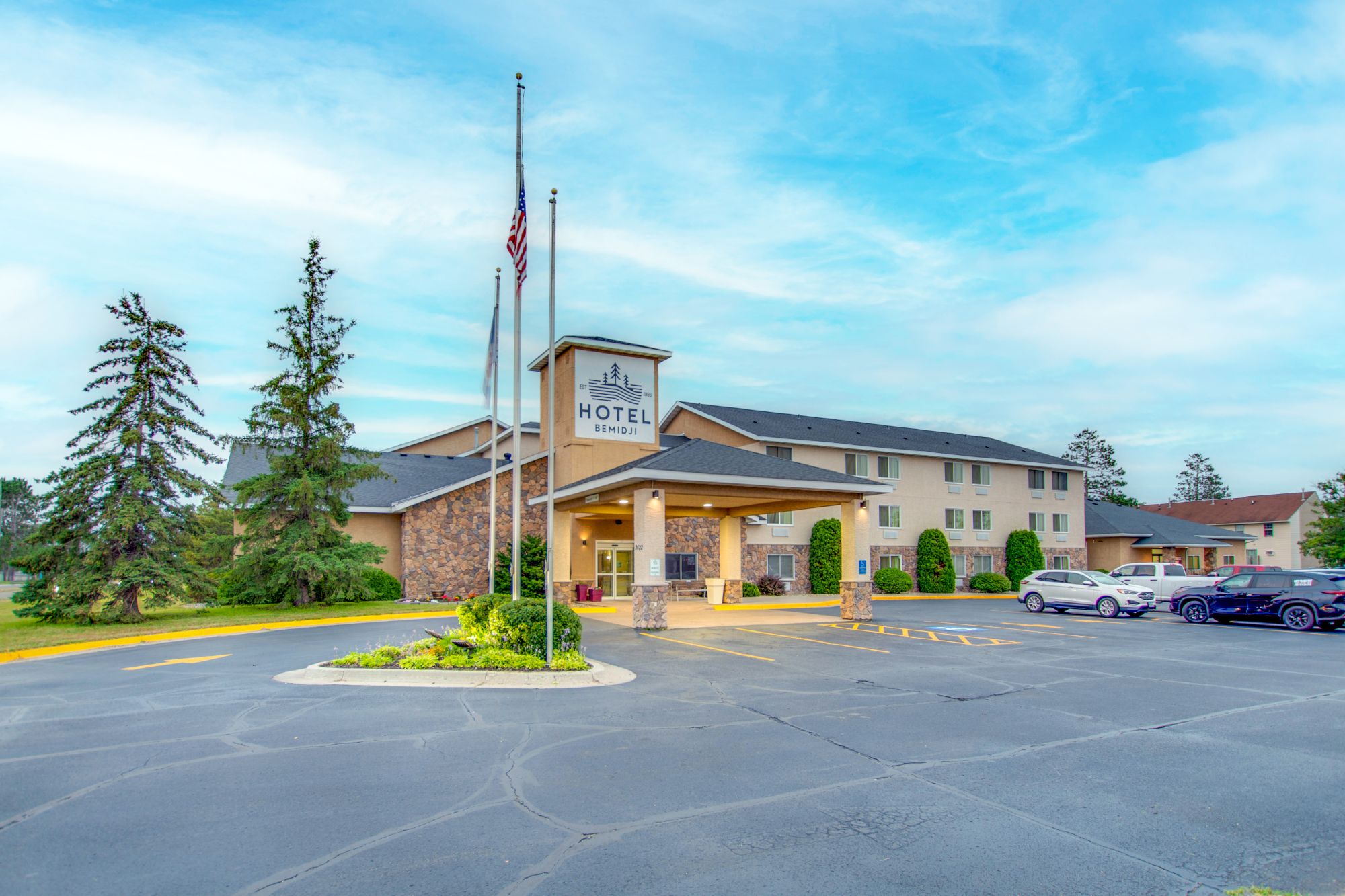 The image shows the exterior of a hotel with a parking lot, flags, and parked cars under a partly cloudy sky.