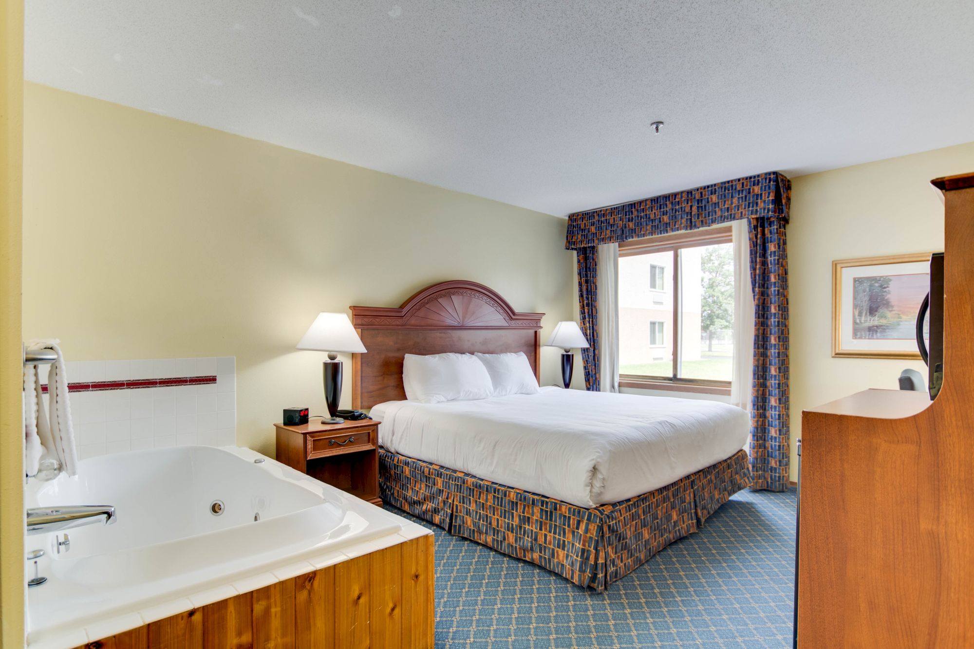 The image shows a hotel room with a king-size bed, two bedside tables with lamps, a window, a bathtub, and a wooden dresser.