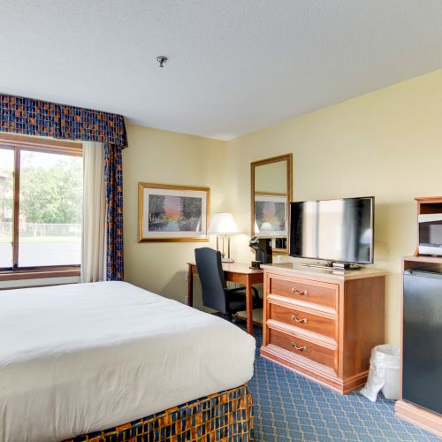 The image shows a well-organized hotel room with a bed, desk, chair, TV, microwave, mini-fridge, and window.