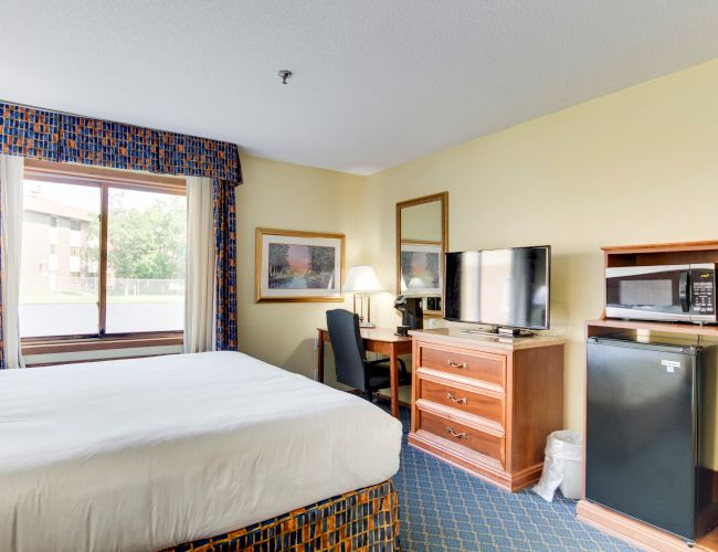 The image shows a well-organized hotel room with a bed, desk, chair, TV, microwave, mini-fridge, and window.