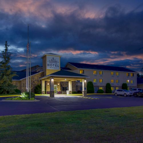 This image shows a hotel at dusk with a lit sign, parking lot, and surrounding trees against a dramatic evening sky.