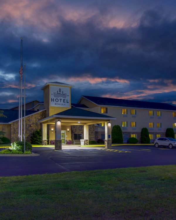 This image shows a hotel at dusk with a lit sign, parking lot, and surrounding trees against a dramatic evening sky.