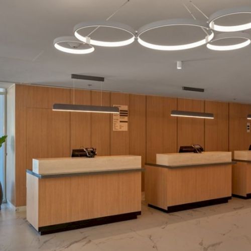 A modern, airy lobby with three wooden reception desks, circular ceiling lights, marble floor, and potted plants near glass doors, welcoming reception.