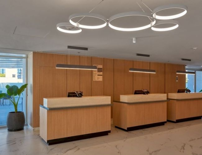 A modern, airy lobby with three wooden reception desks, circular ceiling lights, marble floor, and potted plants near glass doors, welcoming reception.