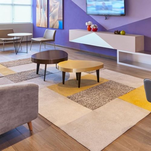 A modern, colorful living lobby with a gray sectional sofa, round coffee tables, lounge chairs, a purple accent wall, and a TV above a white console.