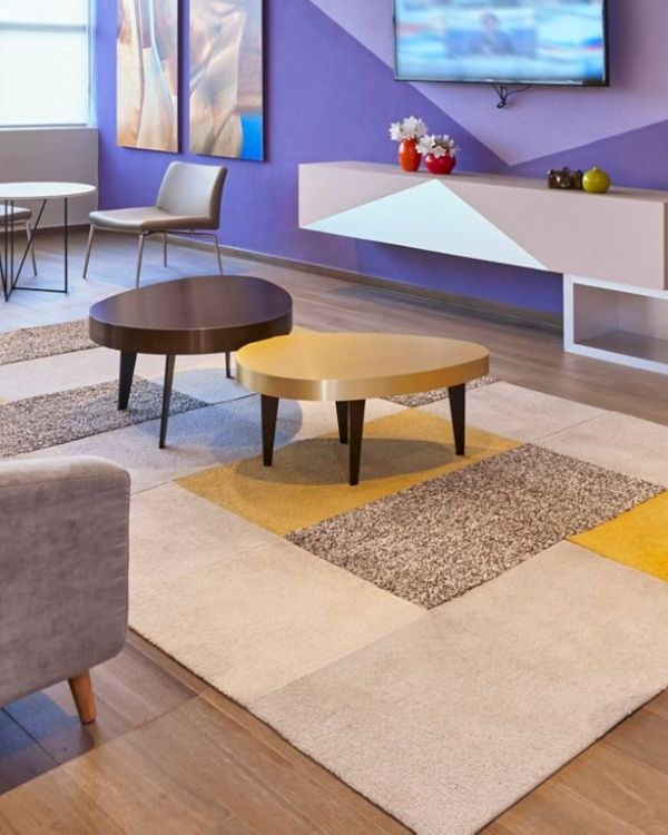 A modern, colorful living lobby with a gray sectional sofa, round coffee tables, lounge chairs, a purple accent wall, and a TV above a white console.