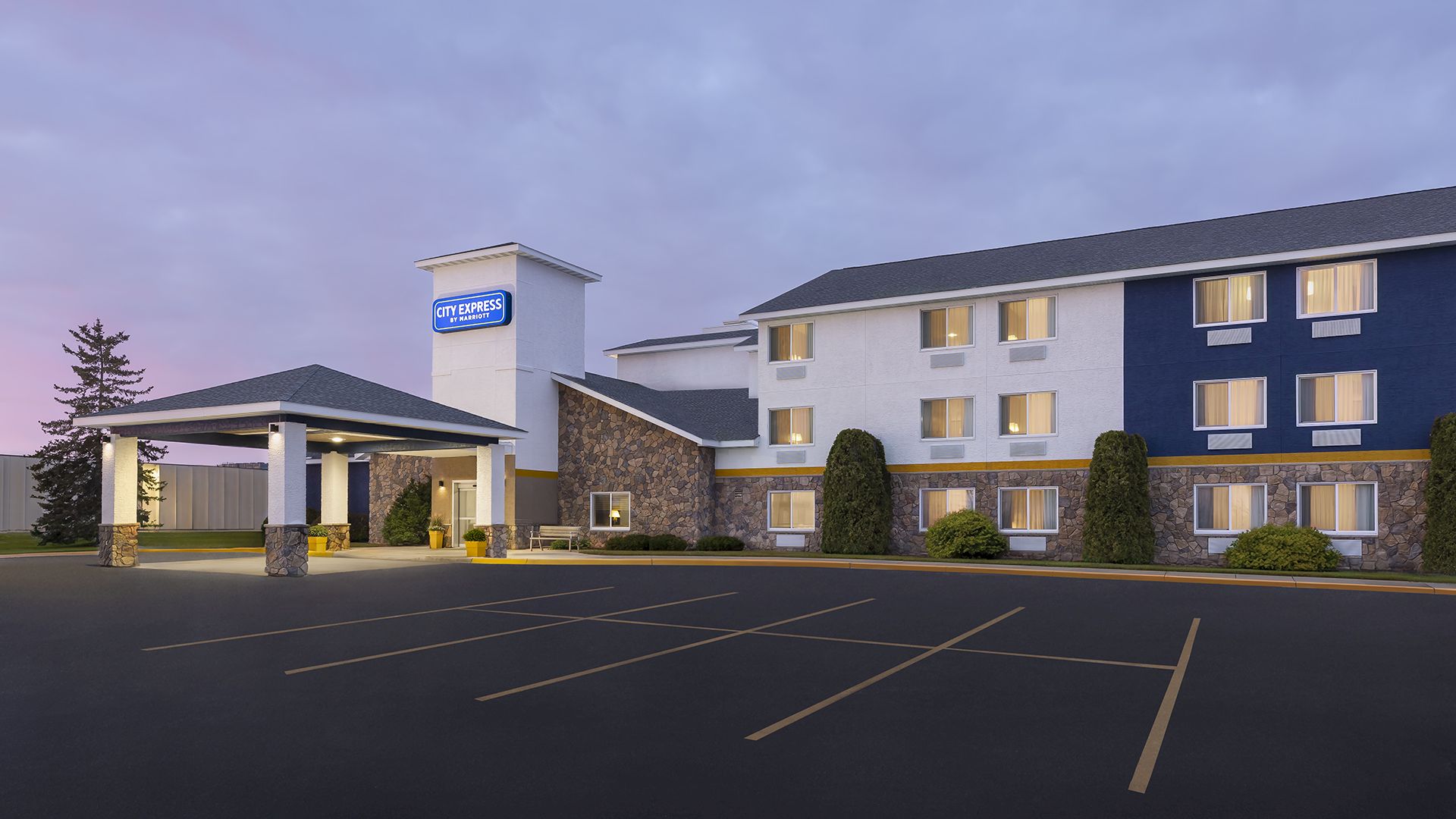 A hotel exterior at dusk with a lit entrance and a large, mostly empty parking lot in front, modern building, few trees.