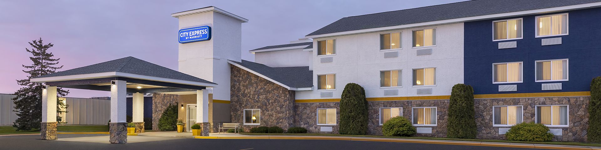A hotel exterior at dusk with a lit entrance and a large, mostly empty parking lot in front, modern building, few trees.