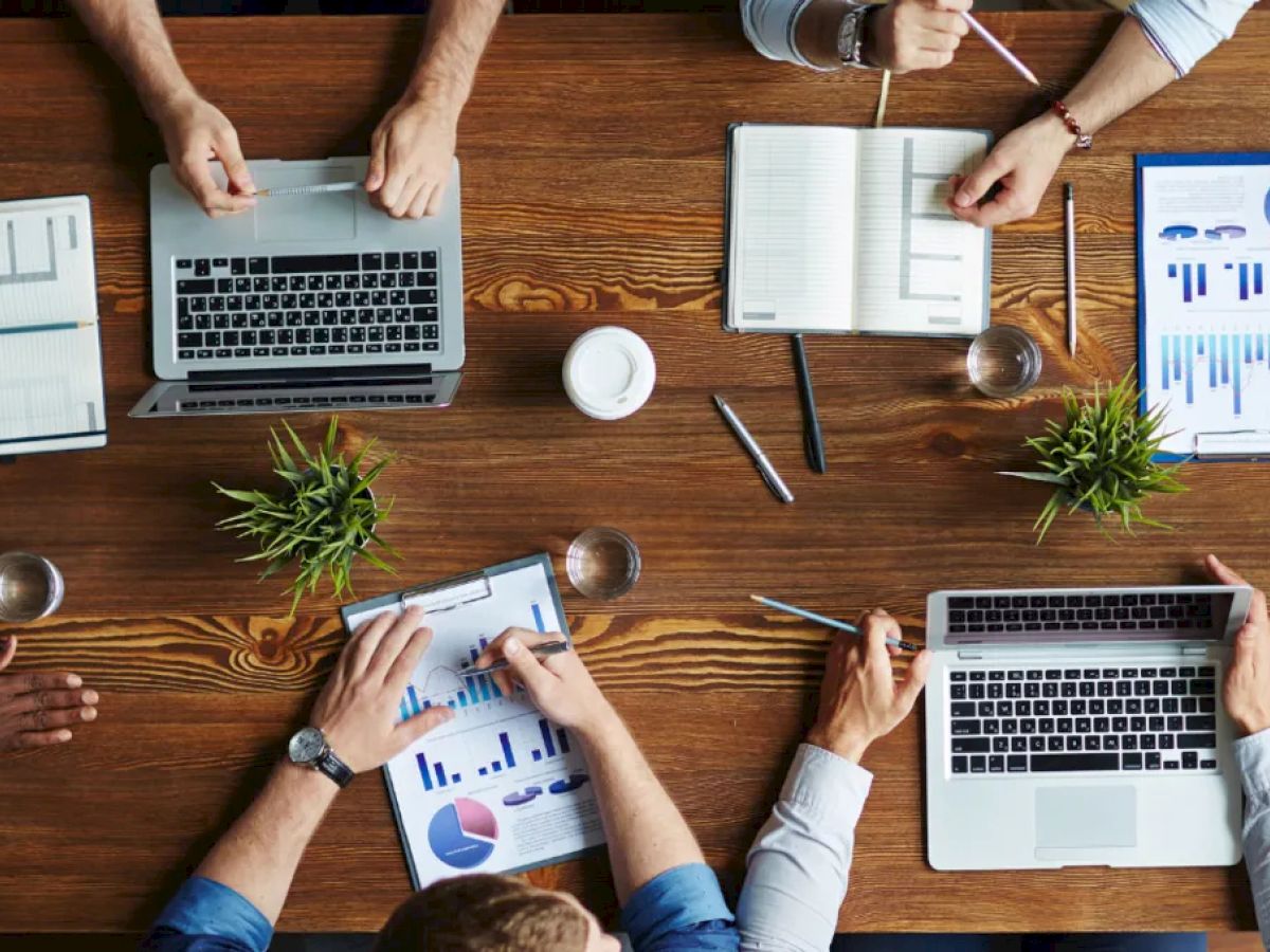 People are working at a wooden table with laptops, tablets, notebooks, and documents showing charts and graphs, with plants and coffee cups.