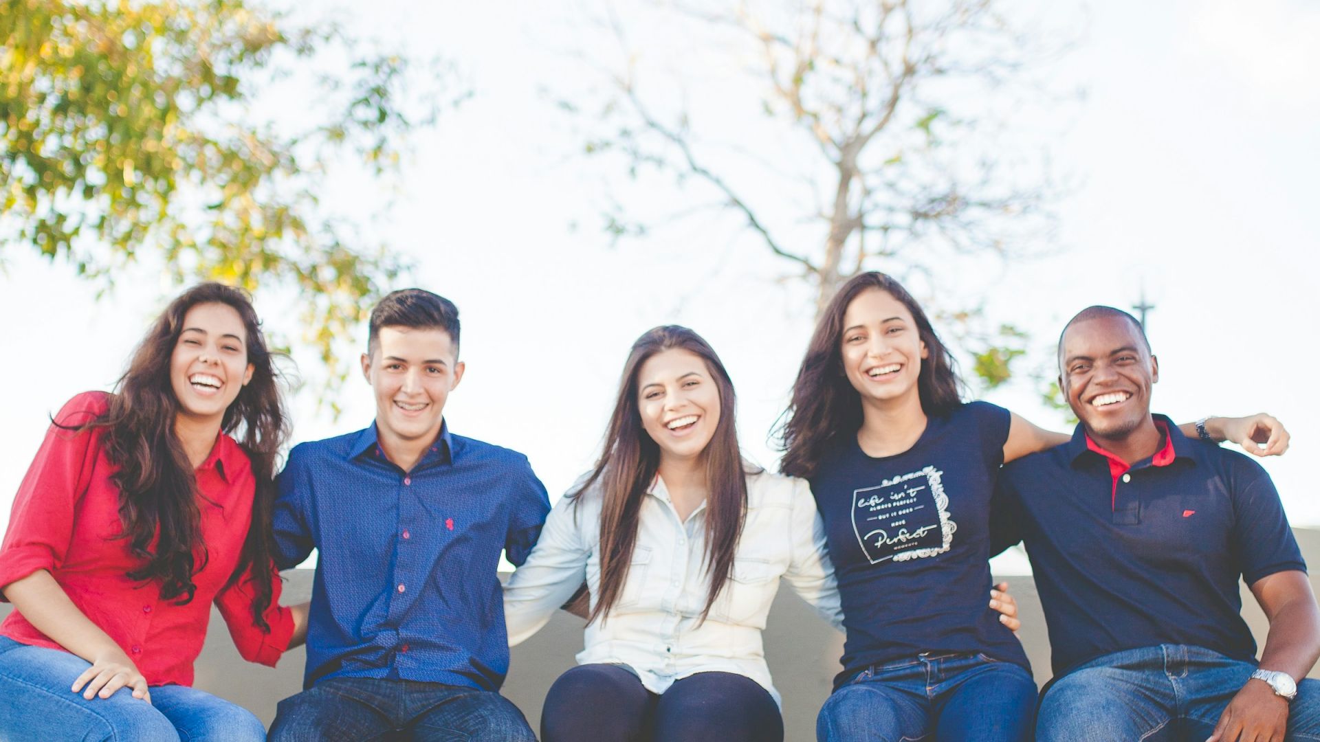 Five people are sitting on a ledge outdoors, all smiling and dressed casually. They seem to be enjoying a sunny day together.