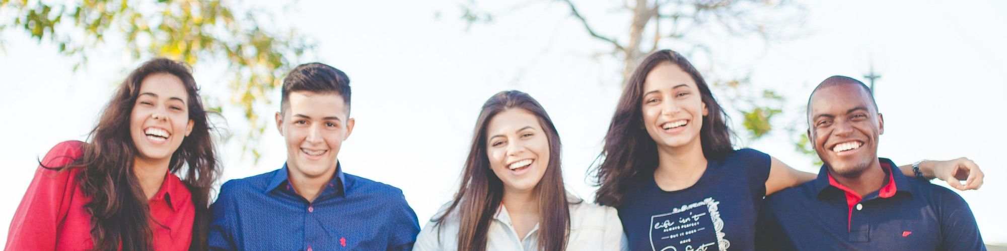 Five people are sitting on a ledge outdoors, all smiling and dressed casually. They seem to be enjoying a sunny day together.