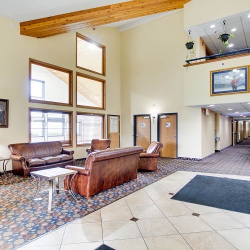 A cozy hotel lobby featuring leather sofas, side tables, wall art, a high ceiling with wooden beams, and a corridor leading to rooms.