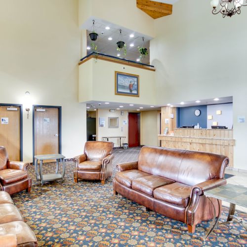 A hotel lobby with leather couches, a reception desk, a chandelier, carpeted floor, and a mezzanine with plants overlooking the space.