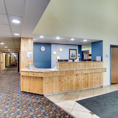 The image shows a hotel lobby with a wooden front desk, a long carpeted hallway, and walls with framed art and lights.