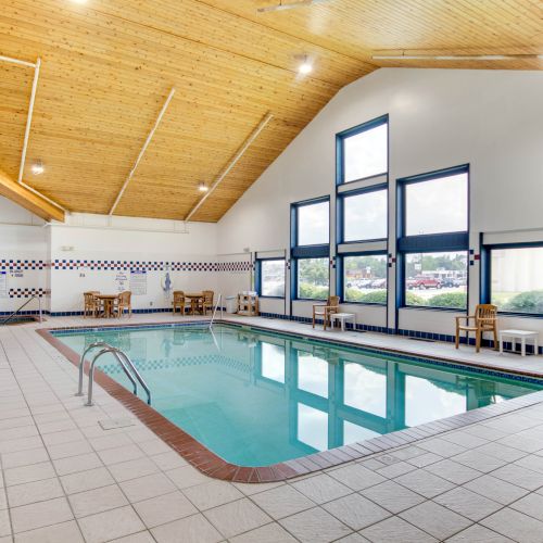 An indoor swimming pool with a tiled deck, lounge chairs, and large windows providing natural light. Wooden ceiling adds warmth to the space.