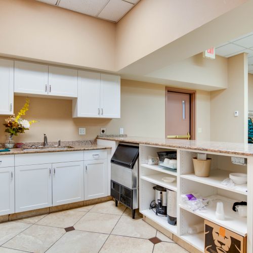 A small kitchen area with white cabinets, granite countertops, and various kitchen items. There's also a room with stacked chairs in the background.