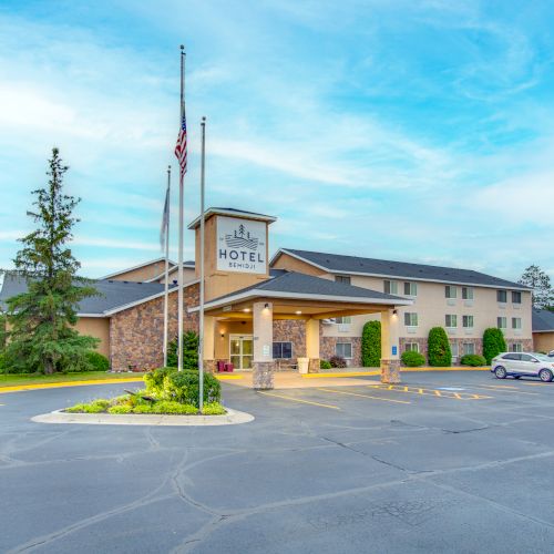 The image shows a hotel building with a clear entrance, surrounded by trees and parked cars, under a blue sky with a few clouds.